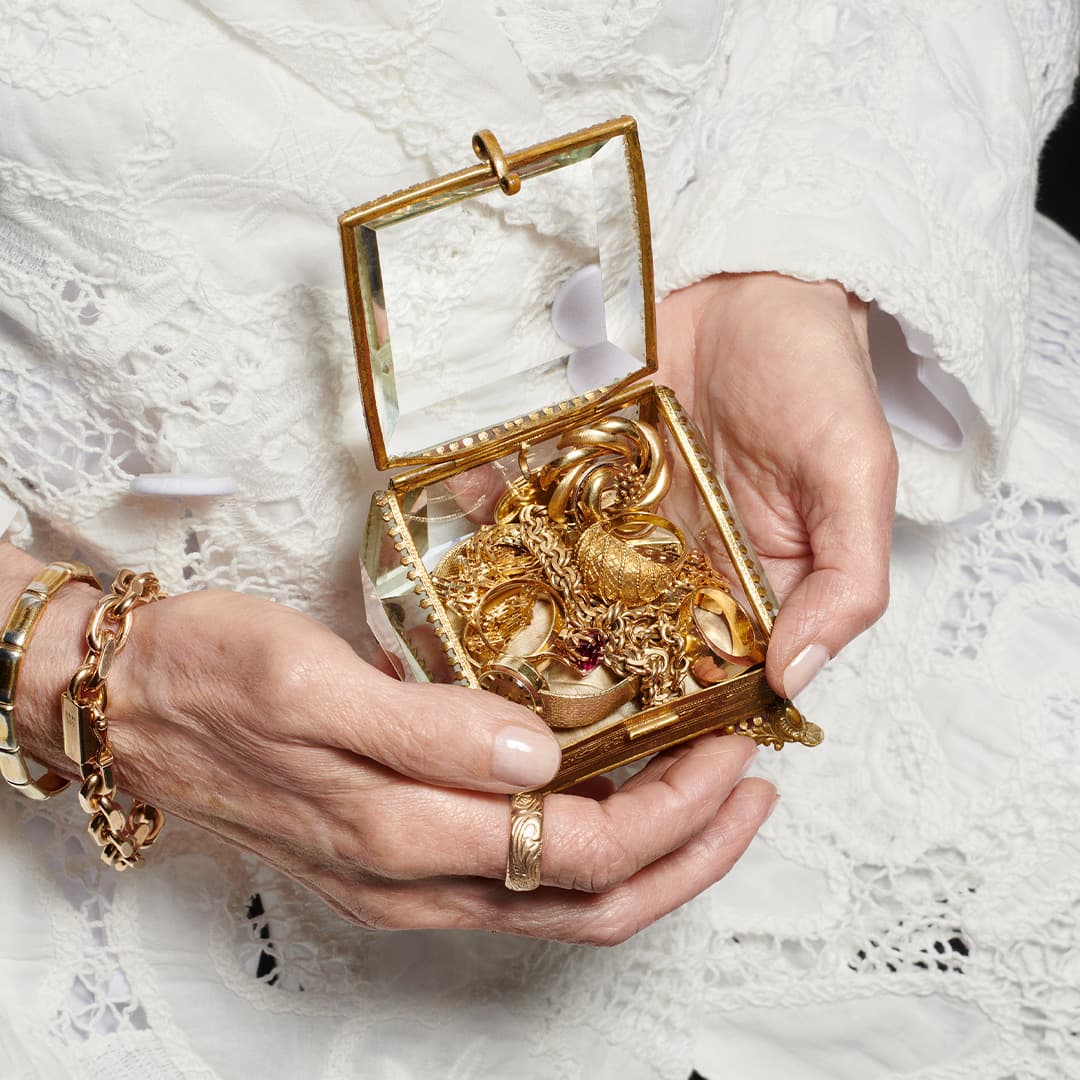Hender i hvitt blondeplagg holder et dekorert glasskrin fylt med gullsmykker. Personen har på gullringer og armbånd.