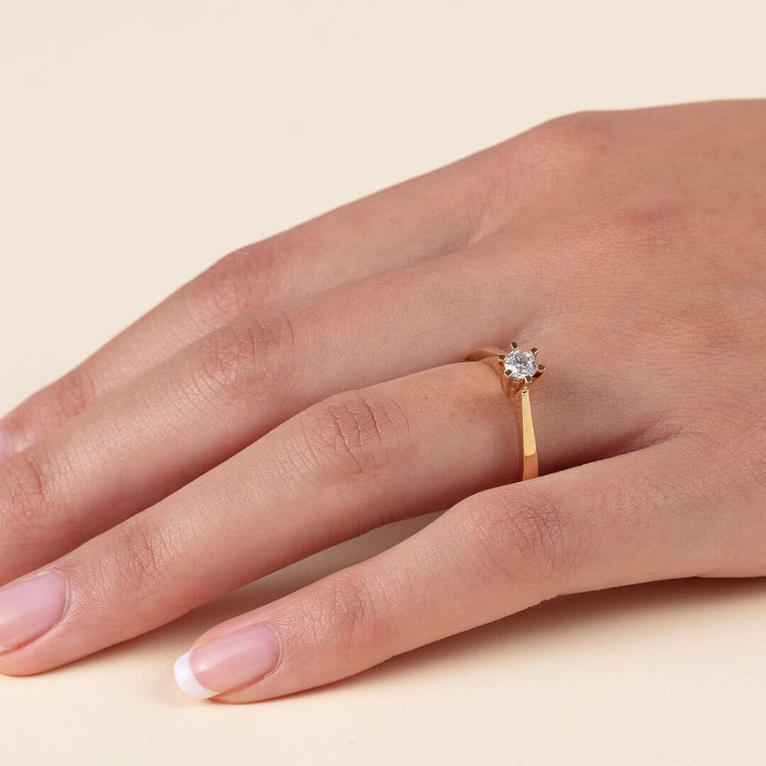 En gullring med en enkelt diamant på en hånd med pent manikyrerte negler mot en beige bakgrunn.