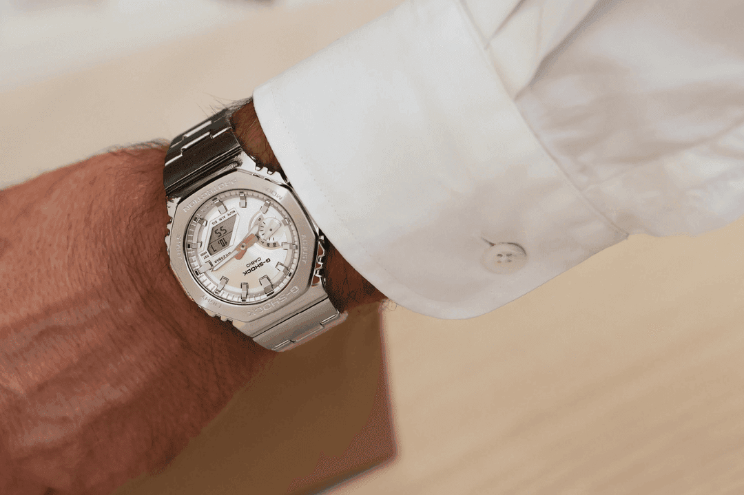 Mann iført et sølvarmbåndsur med lenke i rustfritt stål, hvit urskive og en kneppet hvit skjorte, sett fra et fugleperspektiv.