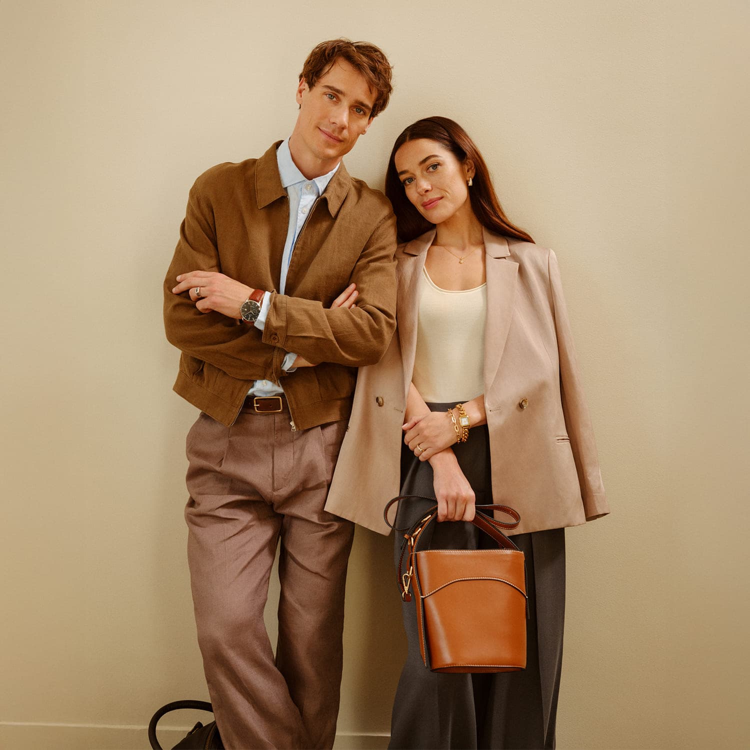 En mann og en kvinne står sammen mot en beige vegg. Begge er stilfullt kledd, og kvinnen holder en brun veske.