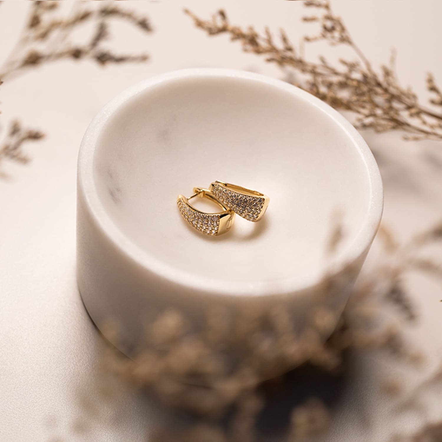 Gullkreoler med små edelstener ligger i en hvit bolle, omgitt av tørkede planter i uskarpt fokus på en glatt overflate.