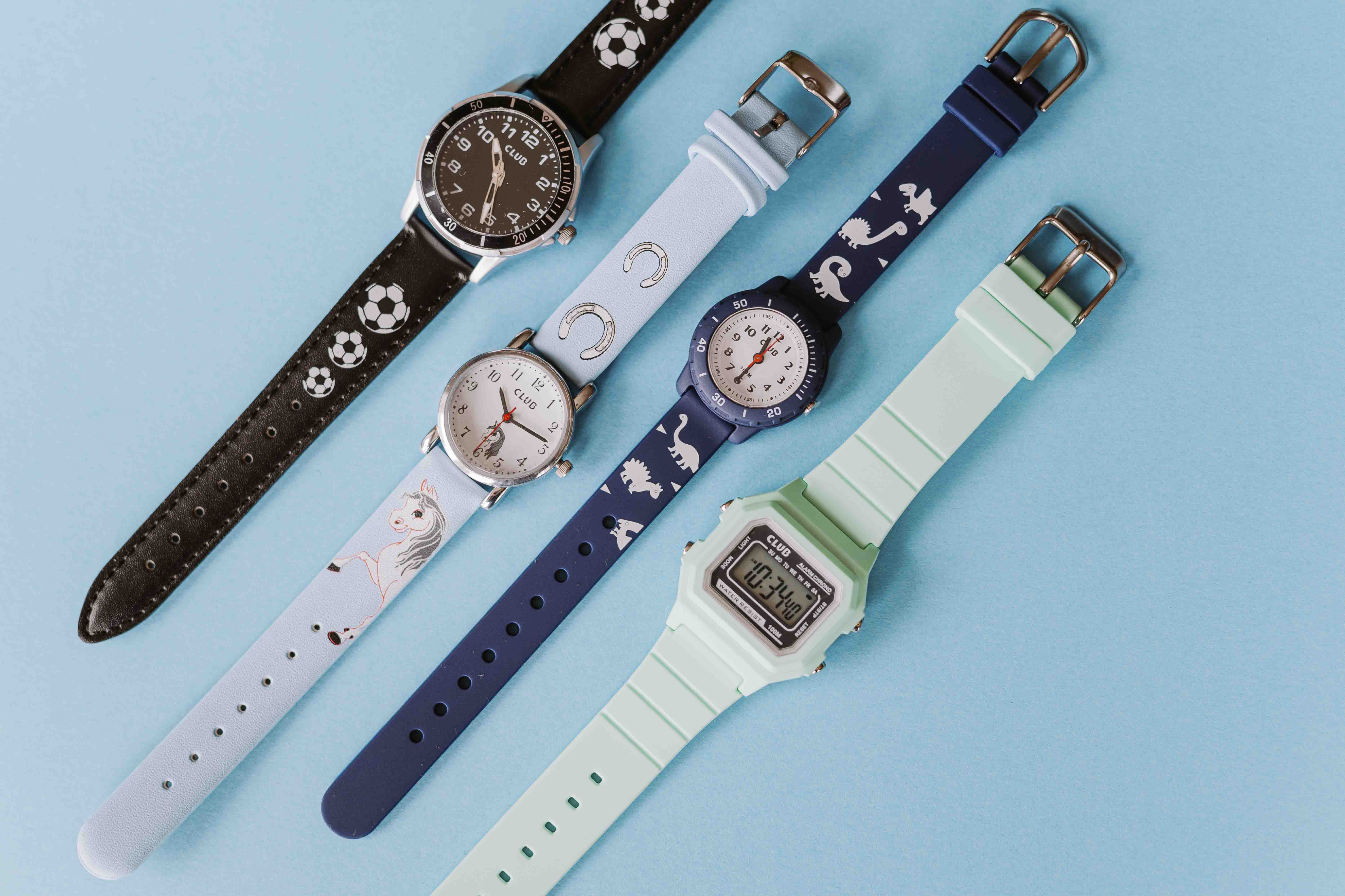 Et lyseblått armbåndsur med hest- og hestesko-design på bunnen. Fem fargerike armbåndsur med forskjellige design, inkludert fotballer, hestesko, enhjørninger og dinosaurer, på en lyseblå bakgrunn, hvilende på en myk beige stoffbakgrunn.