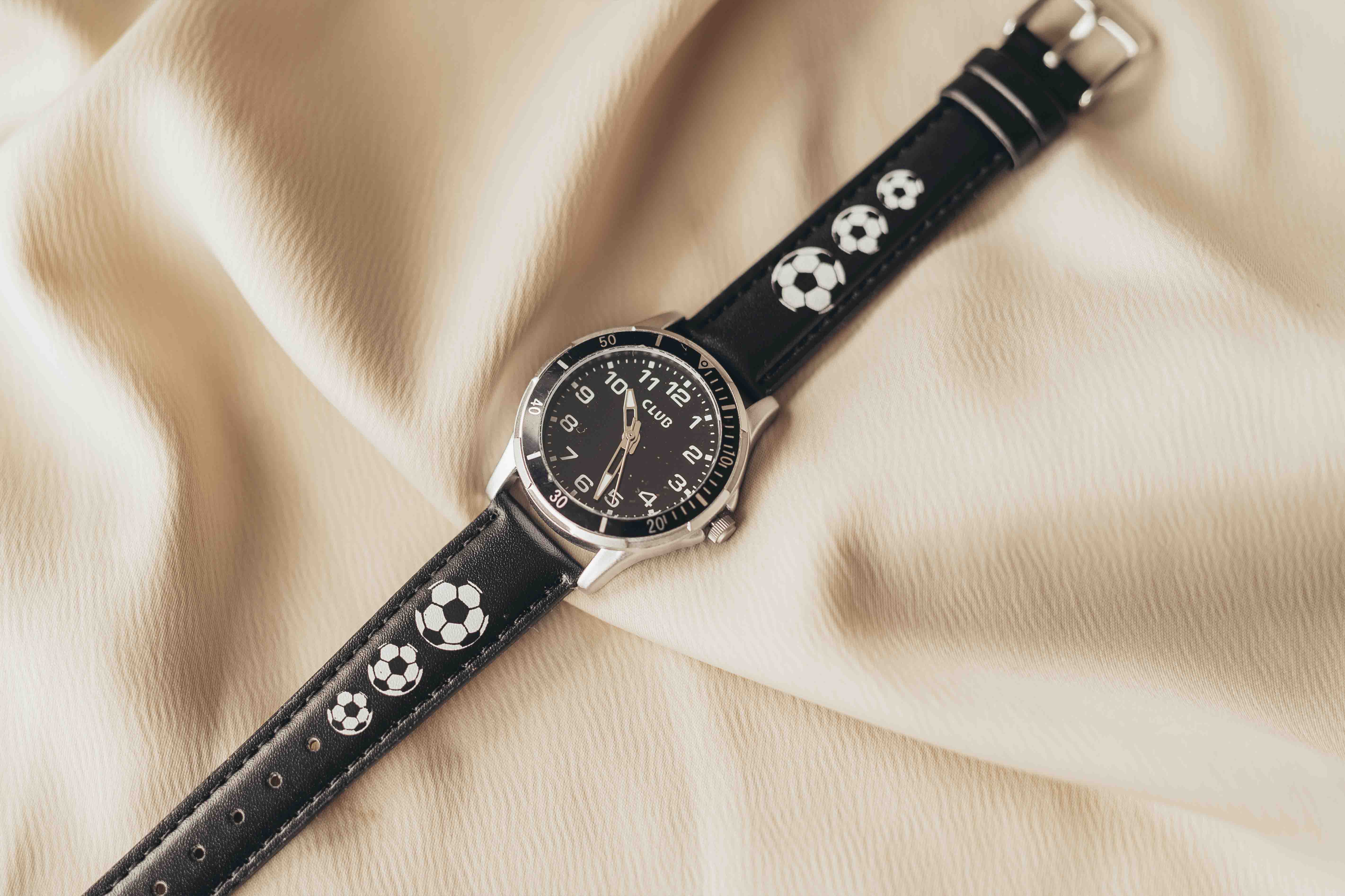 Svart armbåndsur med fotballdesign på remmen, vist på en myk beige stoffbakgrunn.