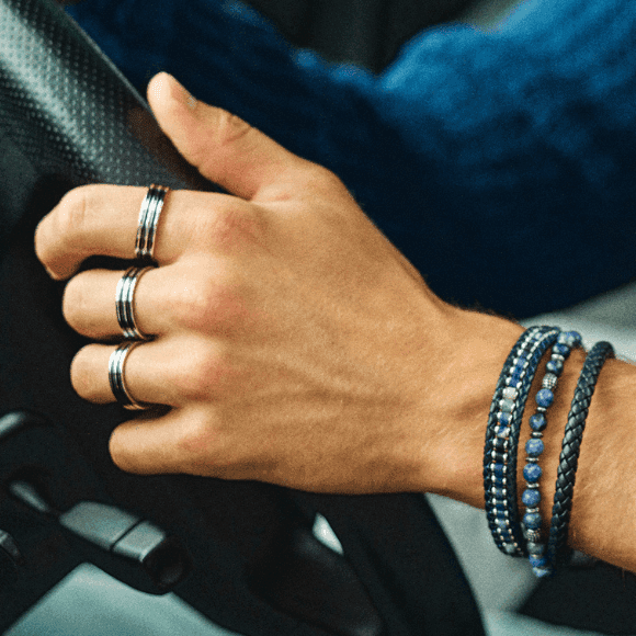 En hånd på et ratt, iført stripete ringer og lagvis sammensatte armbånd i blå- og svarttoner.