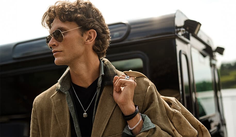 Mann med krøllete hår og solbriller, iført lag-på-lag-halskjeder og en brun jakke, står foran et sort kjøretøy og holder en veske over skulderen.