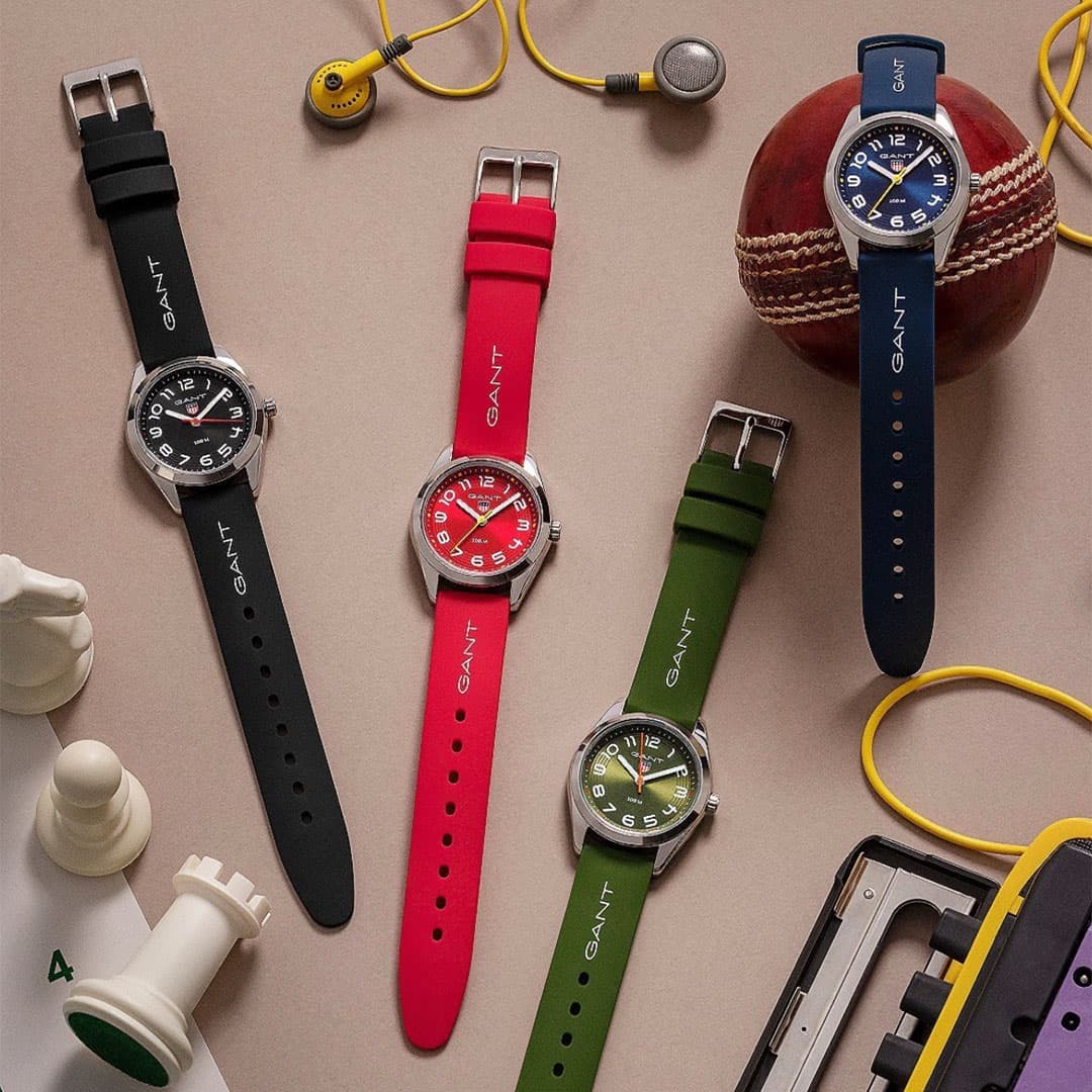 Fire GANT-klokker med fargede remmer (svart, rød, grønn, blå) utstilt blant en cricketball, sjakkbrikker, øretelefoner og dingser.