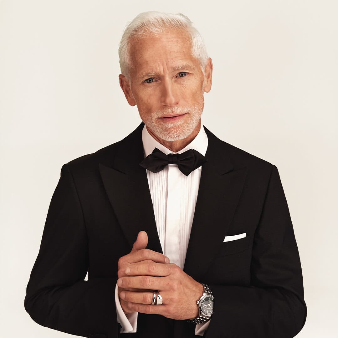 Eldre mann med hvitt hår og skjegg, iført en svart smoking, hvit skjorte og sløyfe, står selvsikkert mot en ensfarget bakgrunn.