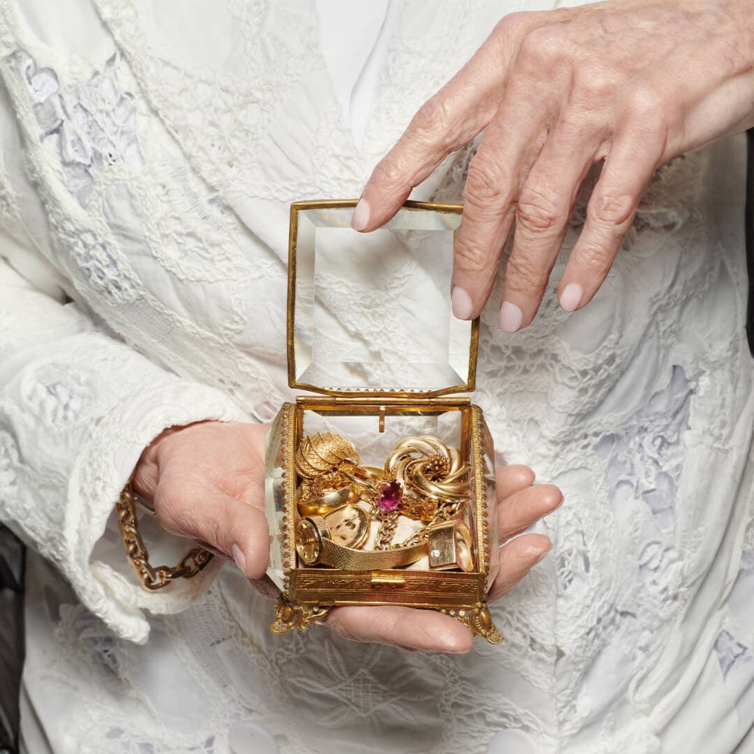 Person i hvitt blondeplagg holder et utsmykket glasskrin med gullsmykker, inkludert et smykke med en rød smykkestein.
