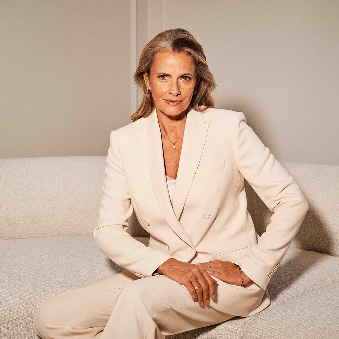 En kvinne i kremhvit dress sitter i en lys sofa mot en enkel bakgrunn og ser selvsikkert direkte inn i kameraet.