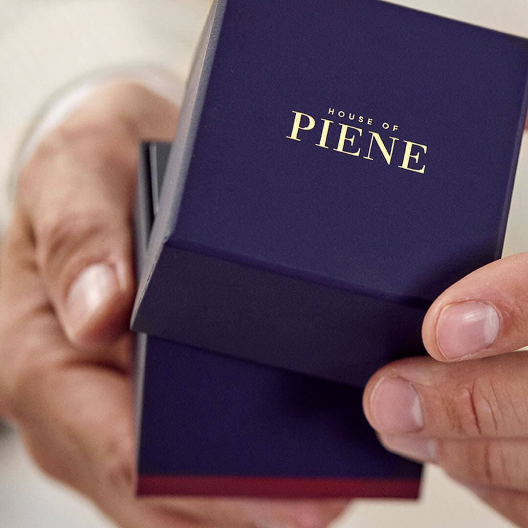 Hender holder en mørkeblå eske merket «House of Piene», som antyder en luksusgave, mot en nøytral, hvit bakgrunn.