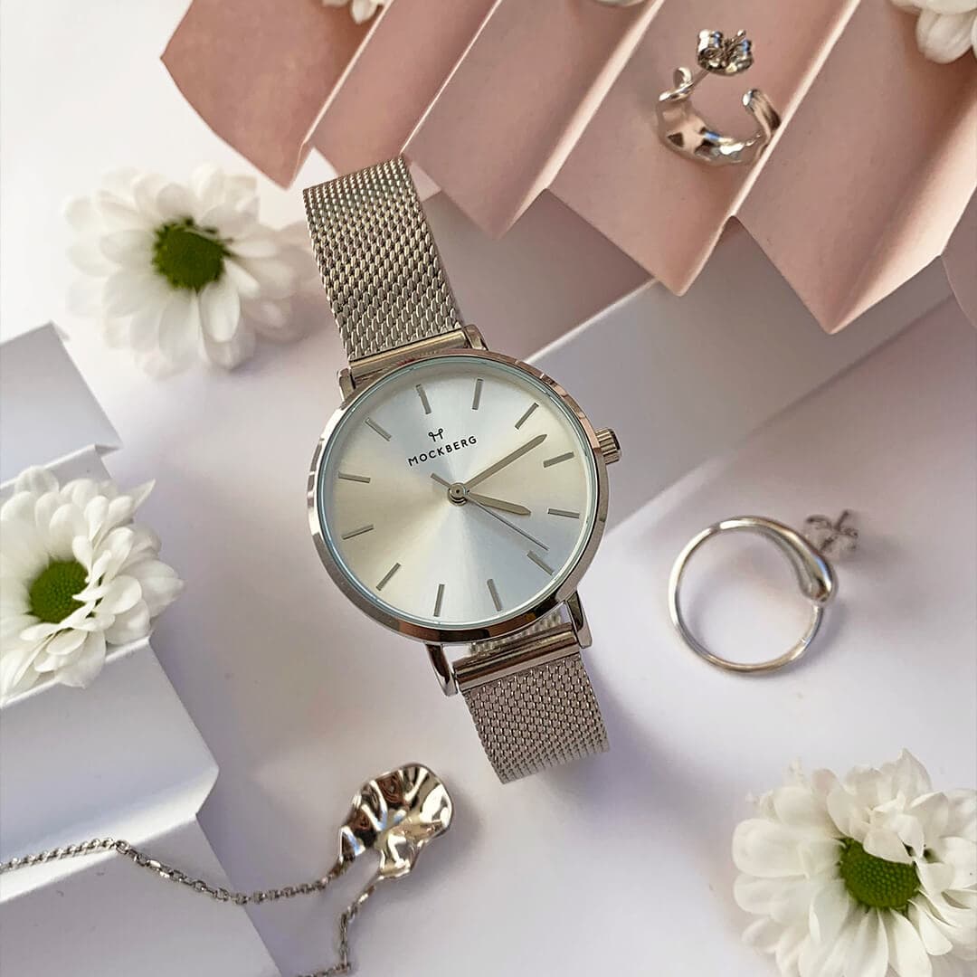 Sølvfarget nettingklokke med hvit urskive på dekorativ bakgrunn, omgitt av tusenfryd og sølvsmykker.