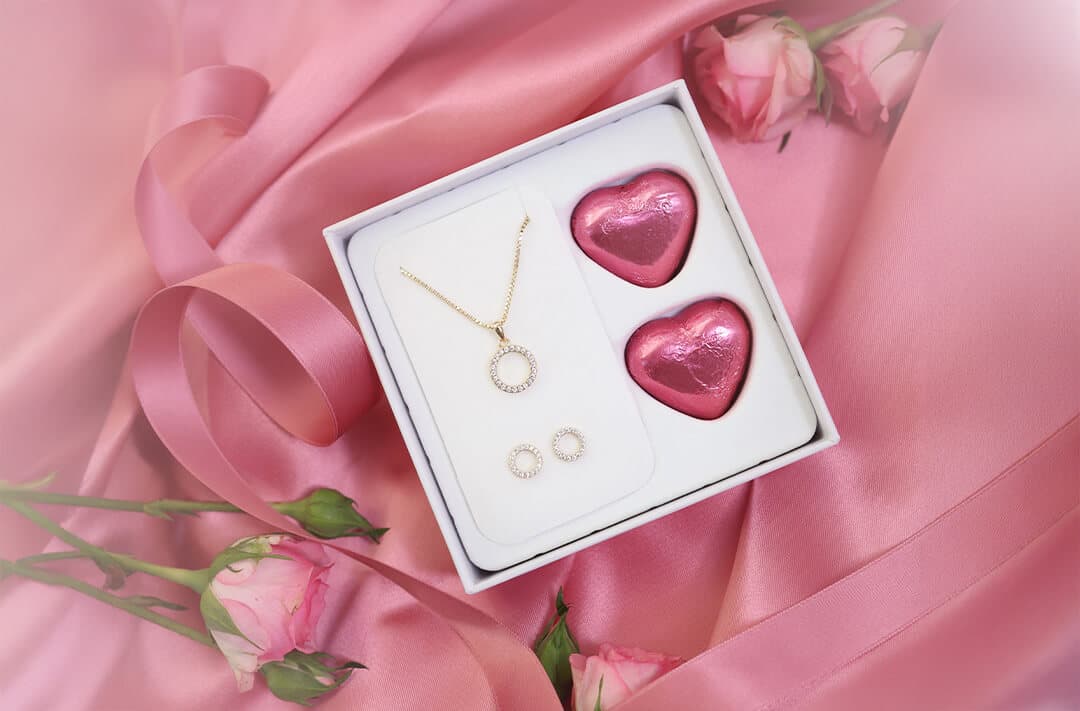 En smykkeeske med et gullkjede og øredobber ved siden av to hjerteformede sjokolader på rosa sateng, omgitt av roser.