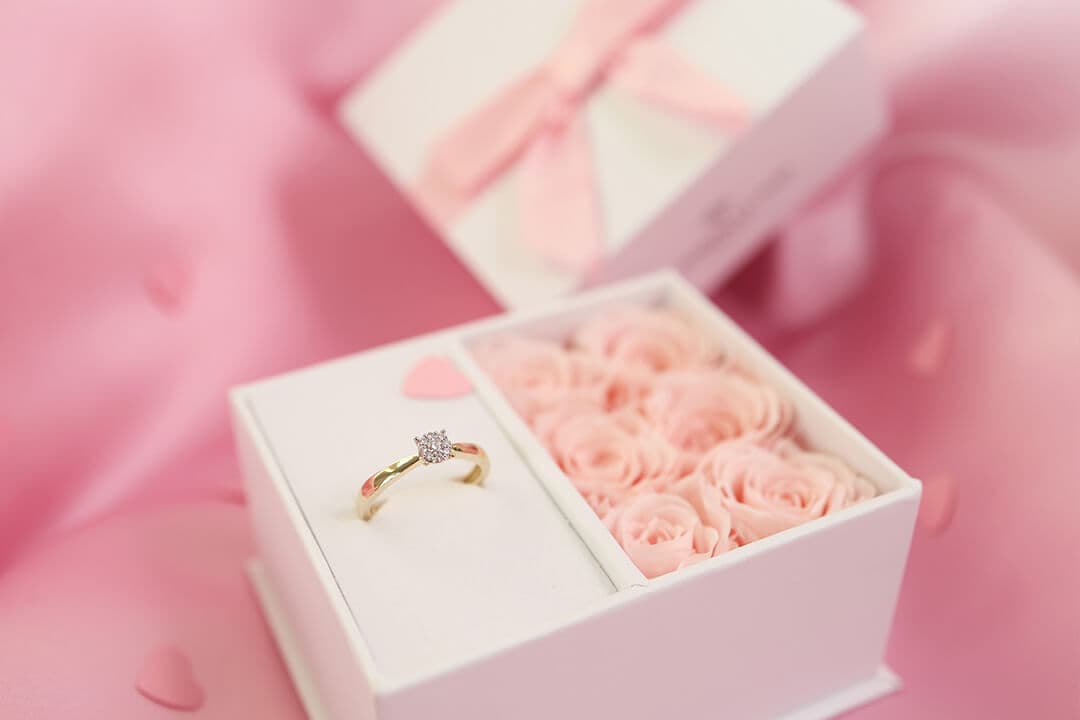 En forlovelsesring i en eske ved siden av rosa roseformede dekorasjoner, på en rosa satengbakgrunn.