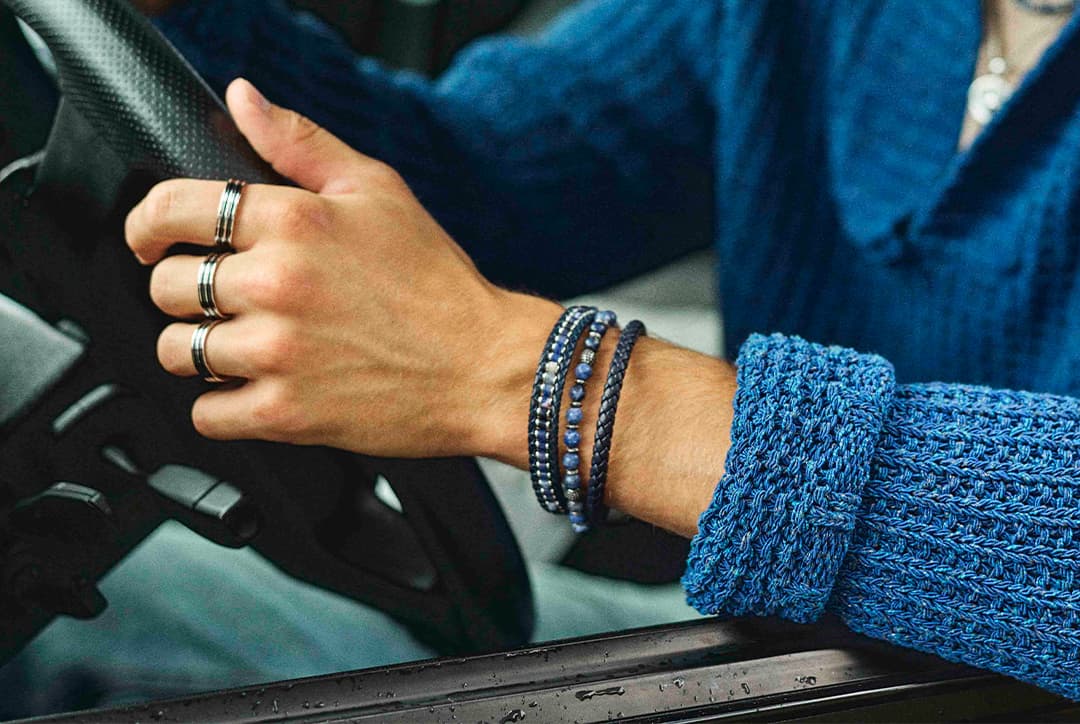 Person med ringer og armbånd som styrer en bil. Blå genser med detaljert strikkemønster.