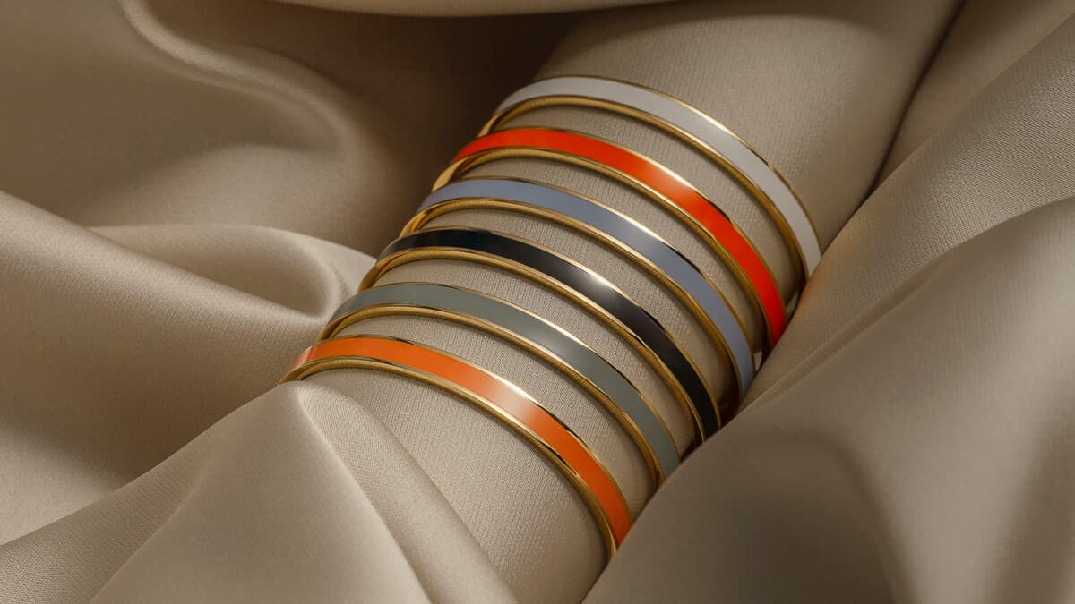 Stabel med armringer i gull og emalje i ulike farger, inkludert oransje og grått, som hviler på mykt, beige stoff.