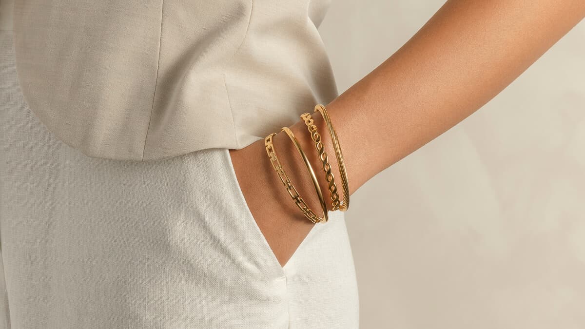 Nærbilde av en persons hånd i en lomme, iført lyse klær og pyntet med tre armringer i gull.