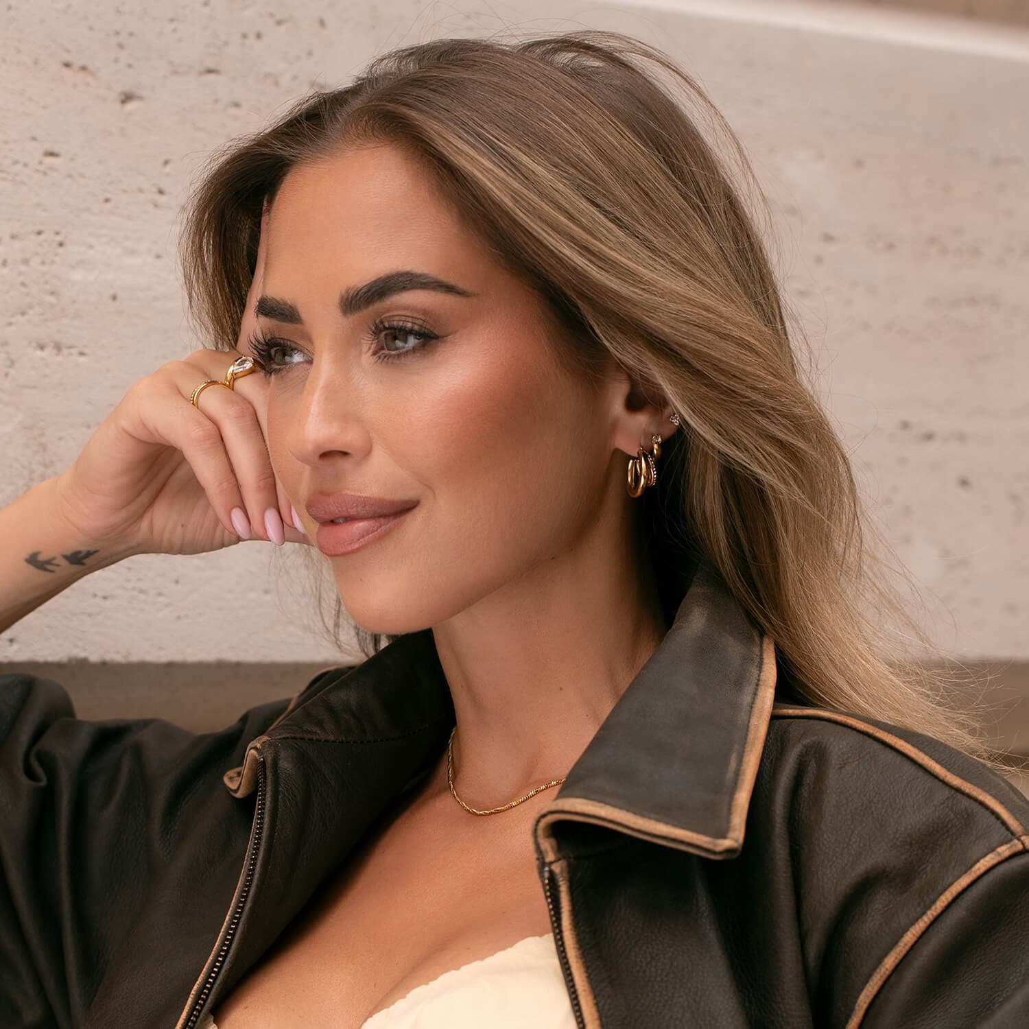 Kvinne med langt hår og gulløreringer, iført skinnjakke, hviler hodet på hånden og ser tankefullt til siden.