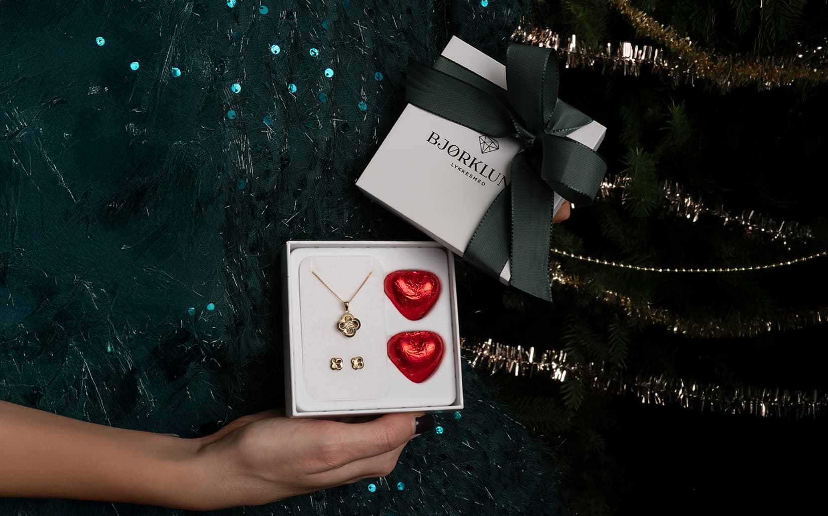 En kvinne holder en gaveeske med et halskjede, øredobber og røde hjerteformede sjokolader, foran et pyntet juletre.