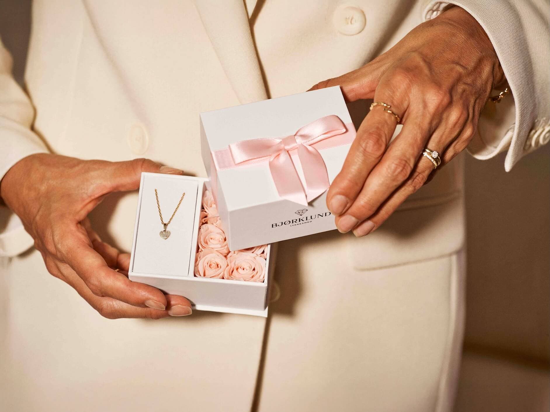 Person i hvit dress som holder en gaveeske med et hjerteanheng og en blomstereske knyttet med et rosa bånd.
