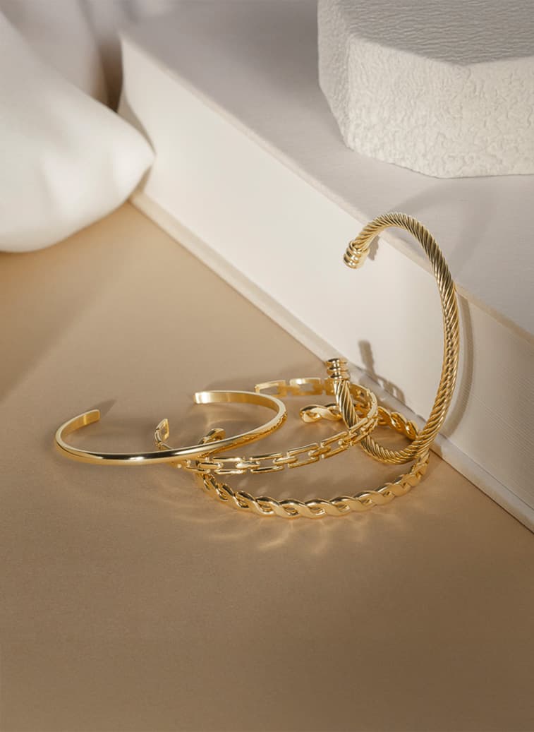 Et sett med gullarmringer, inkludert tvinnede og glatte design, elegant presentert på en beige overflate mot en hvit, teksturert bakgrunn.