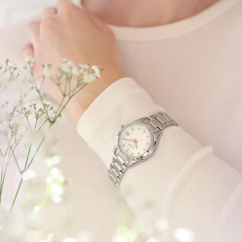 En person med et sølvfarget armbåndsur med rund urskive, hvilende mot en kremfarget skjorte, med delikate hvite blomster i forgrunnen.