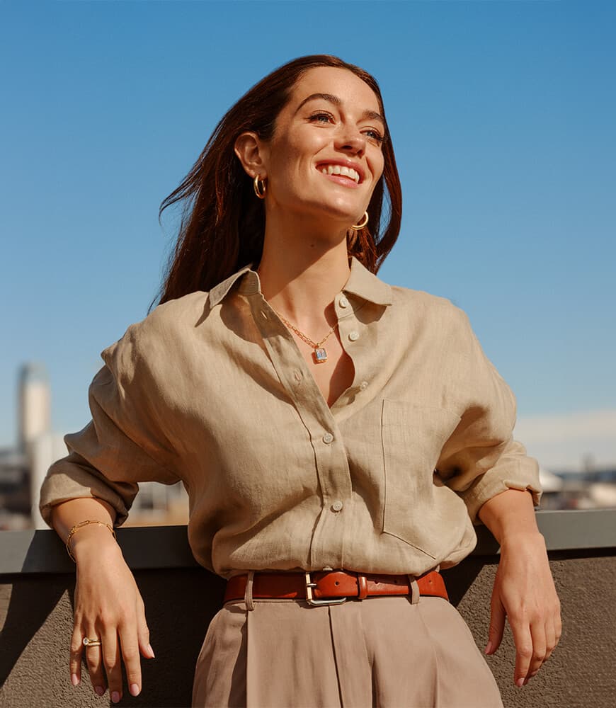 Kvinne som står på et tak, iført en beige skjorte, og smiler lett oppover mot en klar blå himmel i bakgrunnen.