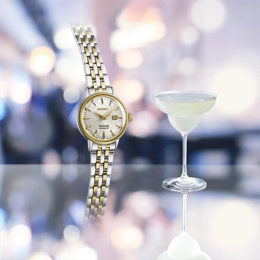 Armbåndsur i gull og sølv med rund urskive vist ved siden av et cocktailglass, mot en uskarp bakgrunn med myke lys.
