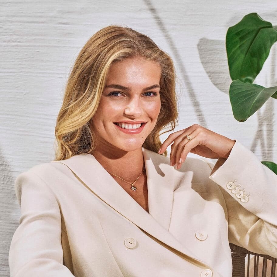 Smilende kvinne med langt blondt hår i en kremfarget blazer, sittende i nærheten av en grønn plante mot en lys teksturert bakgrunn.