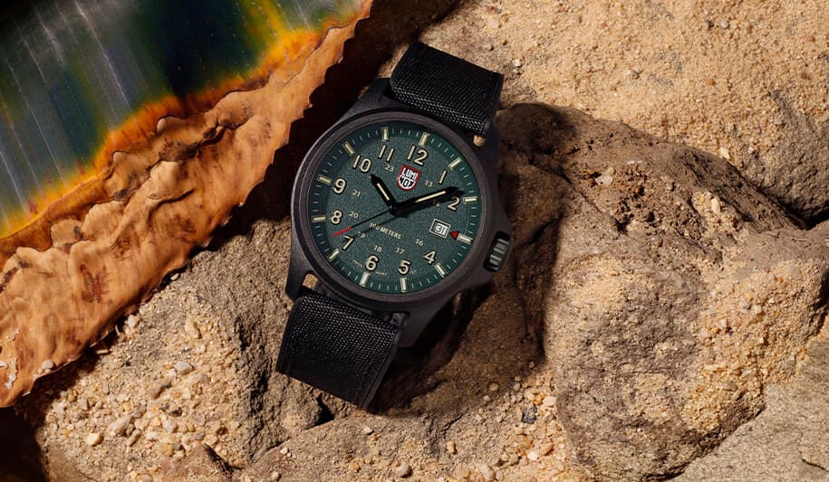 Et armbåndsur med grønn urskive og svart stoffreim hviler på sandsteiner, akkompagnert av en fargerik skjellkant i bakgrunnen.